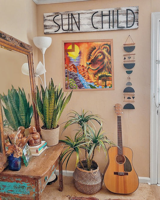 Framed art print with the text 'SUN CHILD' on a wooden background, displayed above a guitar and a wooden cabinet, with decorative items and plants in the room.